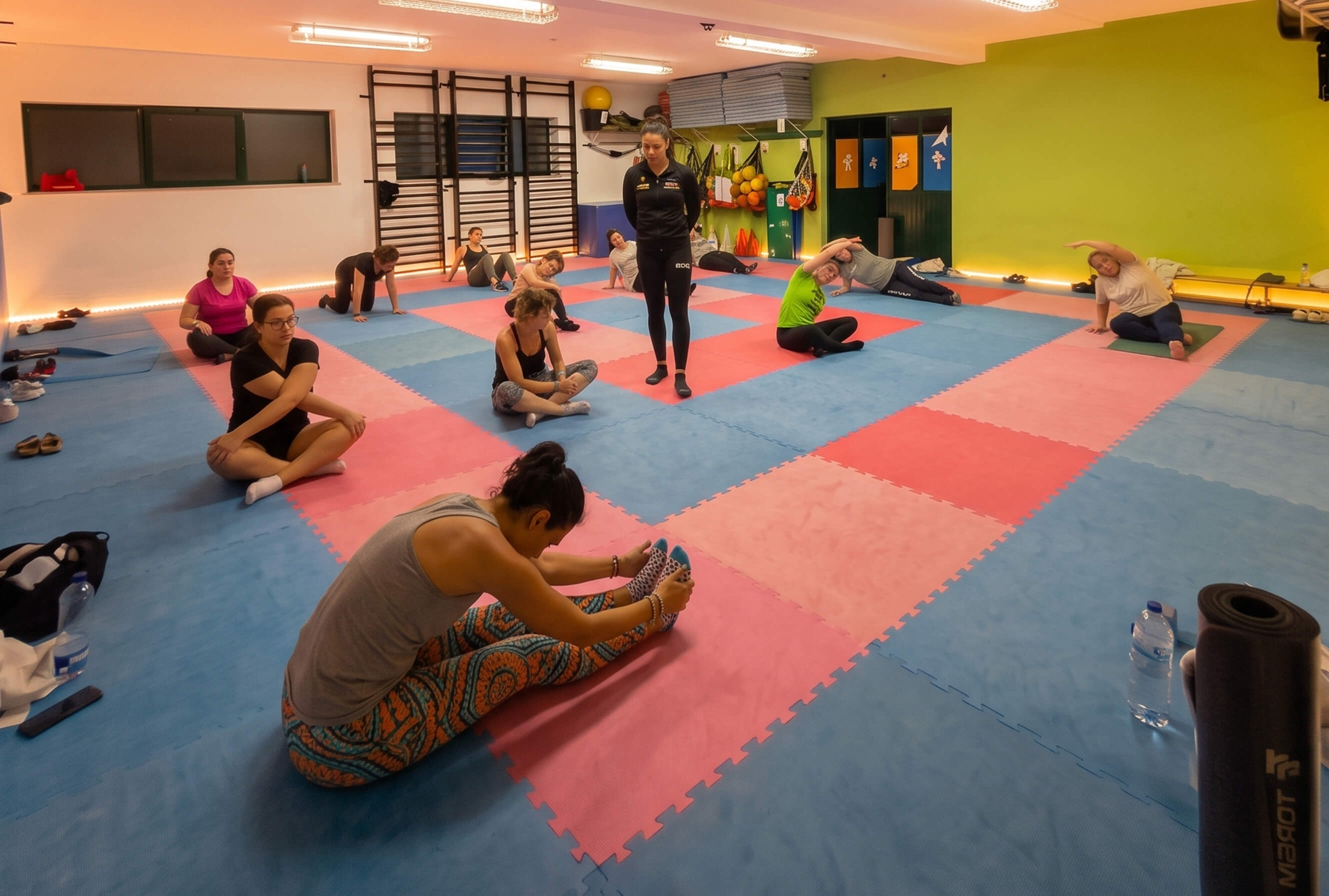 Aula indoor O2Fit em tapetes coloridos, com a treinadora ao centro, na Parede